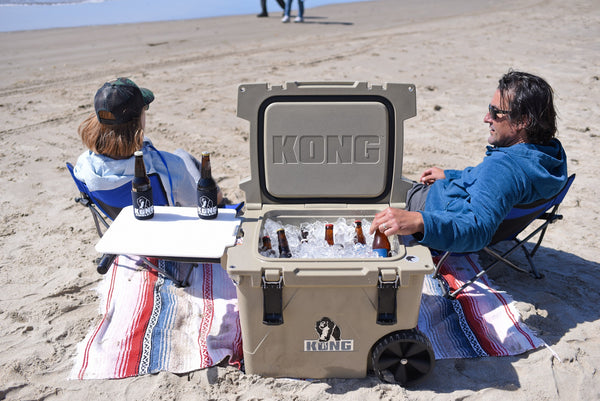 KONG Cruiser 50 quart Cooler on a beach with two people sitting in lawn chairs. Cooler with wheels on a beach