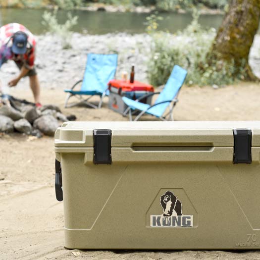 KONG Cooler 70 quart with lid closed. Man starting a camp fire behind with two lawn chairs to the side of the fire